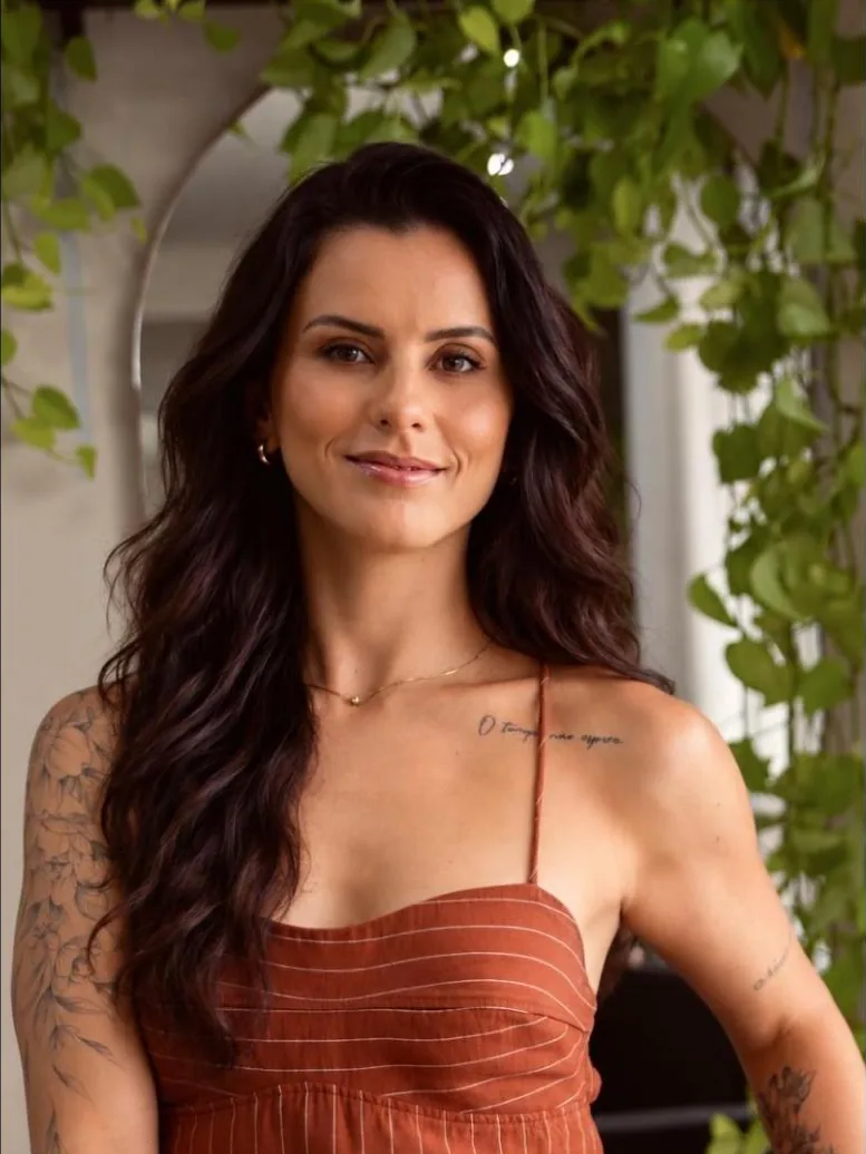 Isa Guido, cabeleireira, de vestido terracota, em frente ao espelho em arco com plantas do salão